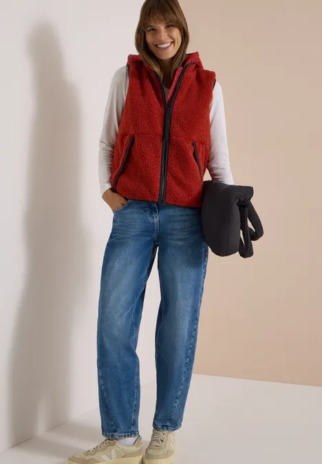 Eine lächelnde Frau trägt rote Weste, weiße Bluse, blaue Jeans und weiße Sneaker, hält schwarzen Mantel.