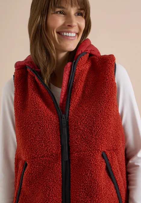 Lächelnde Frau in roter Fleece-Weste mit Kapuze und Reißverschlusstaschen über weißem Langarmshirt.