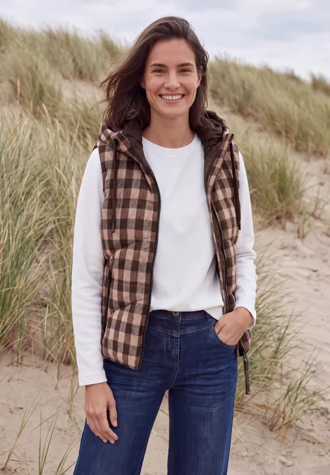 Eine lächelnde Frau steht in den Dünen, trägt ein weißes Longsleeve, eine karierte Weste und Jeans.