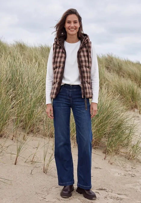 Eine lächelnde Frau trägt Jeans, weißen Pullover und kariertes Gilet am sandigen Dünenstrand.