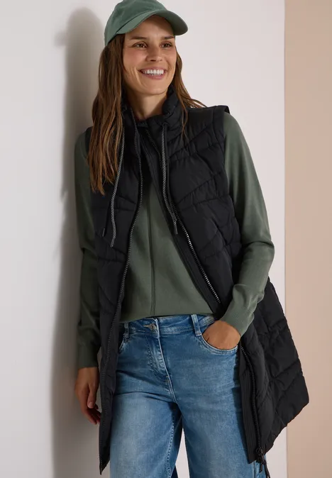Femme souriante en casquette verte, pull kaki, jean bleu et longue doudoune sans manches noire.