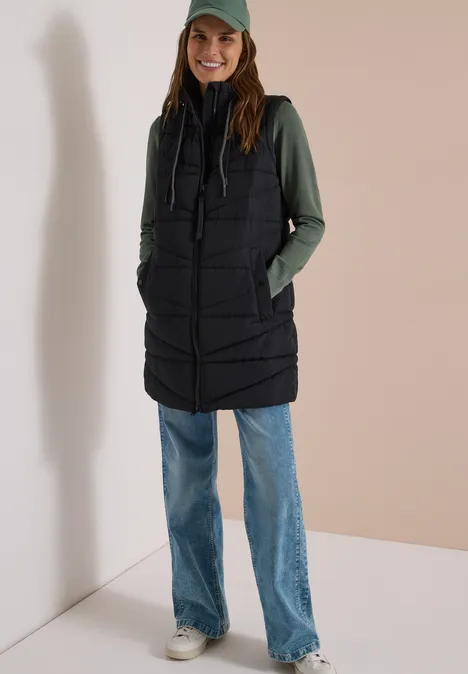 Femme portant une longue veste matelassée noire, un sweat à capuche vert, un jean large et une casquette.