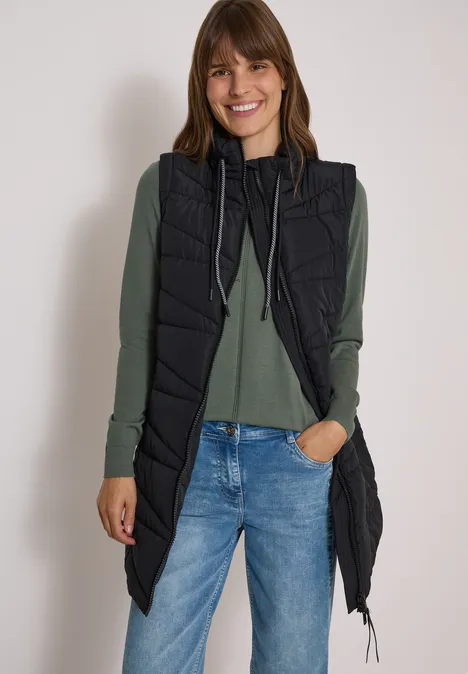 Femme portant une longue doudoune sans manches noire, pull vert et jean bleu, souriante sur fond blanc.