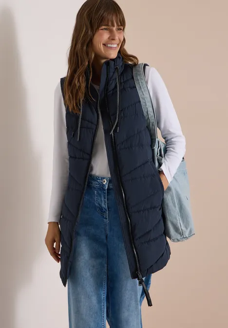 Femme en t-shirt blanc, longue doudoune sans manches bleu marine, jean et sac en bandoulière.