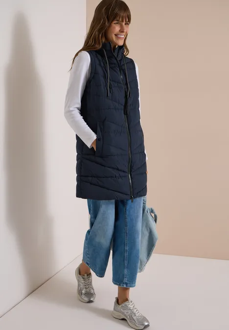 Femme en doudoune sans manches bleu marine, top blanc, jean large, baskets argentées, tenant un sac.