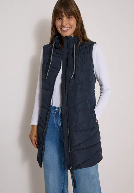 Femme souriante en chemise blanche et jean bleu, portant une longue doudoune sans manches bleu marine.