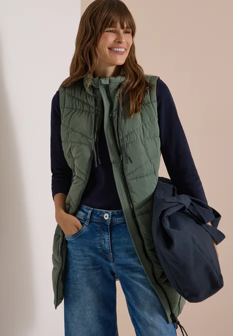 Femme portant une longue veste matelassée verte sur un pull foncé et un jean, tenant une veste bleu marine.