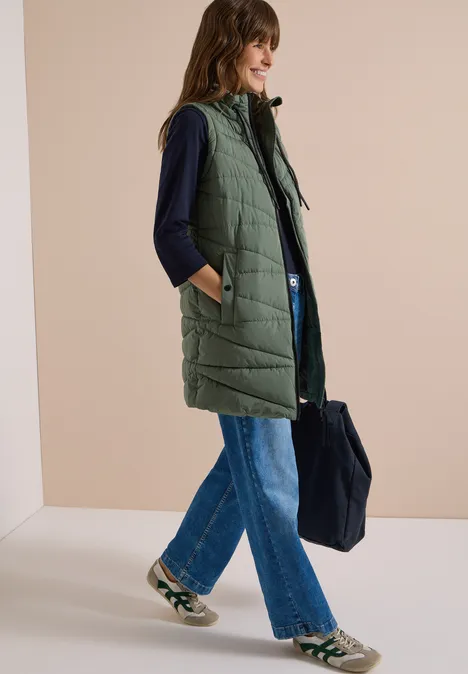 Femme en longue veste matelassée à capuche, top sombre, jean large, baskets et grand sac fourre-tout.