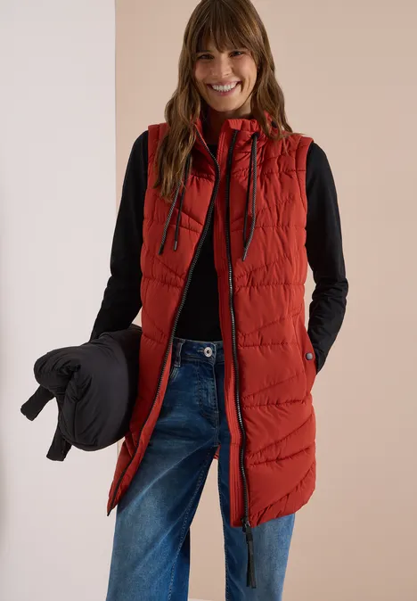 Femme souriante portant une veste sans manches rouge, un top noir et un jean, tenant une doudoune noire.