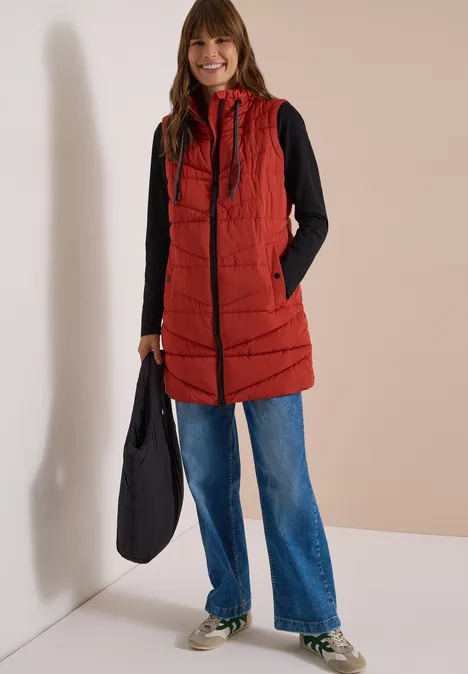 Femme souriante en doudoune longue rouge à capuche, pull noir, jean bleu et sac noir.
