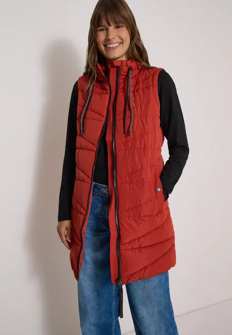 Femme souriante portant une longue doudoune rouge à capuche, un haut noir et un jean ample devant un mur clair.
