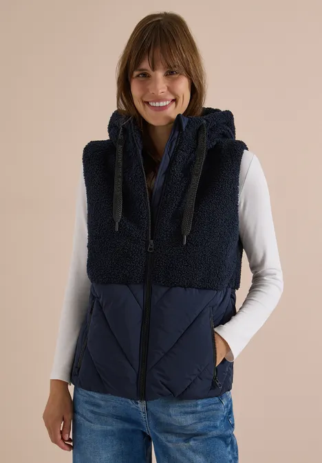 Femme souriante en gilet sans manches bleu marine à capuche polaire, t-shirt à manches longues blanc et jean.