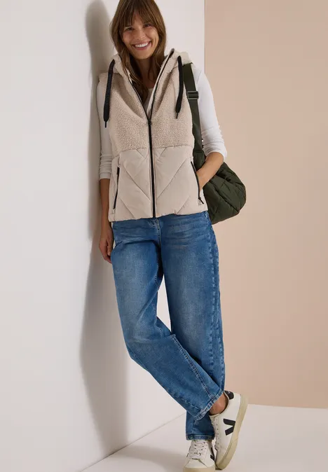 Une jeune femme souriante appuyée contre un mur, en jean, gilet, baskets et sac à dos.