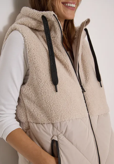 Une femme porte une veste sans manches en sherpa beige avec capuche et poches zippées.