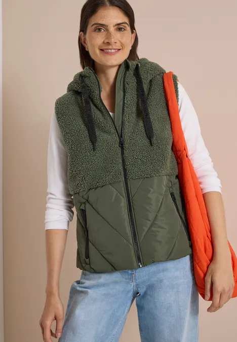 Une femme porte une doudoune sans manches olive à capuche, un tee-shirt blanc à manches longues et un tote bag orange.