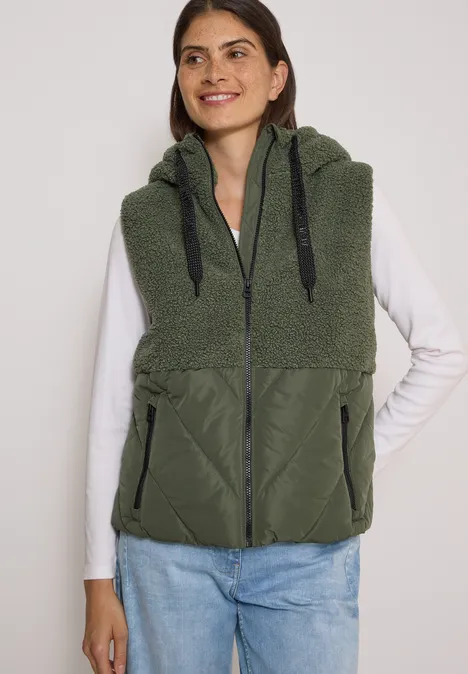 Femme en t-shirt manches longues blanc et jeans porte une veste sans manches en polaire verte à capuche zippée.