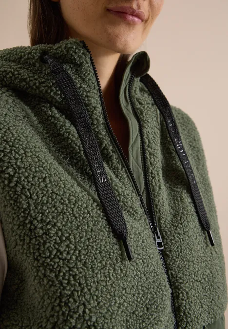 Gros plan d'une personne portant un sweat à capuche teddy vert olive avec zip et cordons noirs.