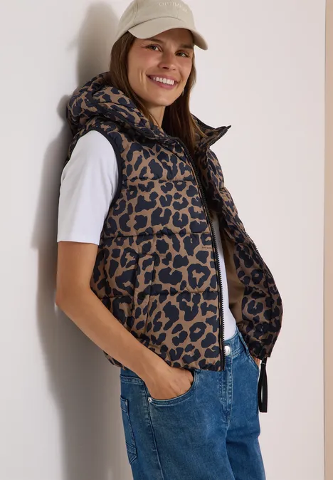 Gewatteerd vest met capuchon en zakken creamy mocha Een glimlachende vrouw draagt een gewatteerd jasje met luipaardprint, een wit T-shirt en een spijkerbroek.