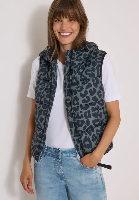 Gewatteerd vest met capuchon en zakken dried olive green Vrouw lacht en draagt een capuchonbodywarmer met dessin op een wit t-shirt en jeans.