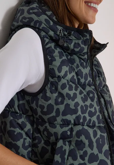 Gewatteerd vest met capuchon en zakken dried olive green Vrouw opzij gefotografeerd met witte longsleeve en gewatteerd, luipaardprint bodywarmer.