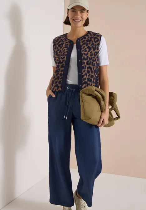 Frau im Freizeitlook mit Cap, weißem T-Shirt, Leo-Weste, weiten Jeanshosen und grüner Umhängetasche.