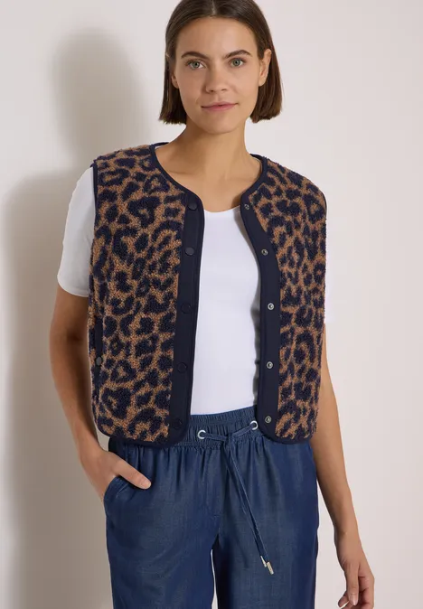 Frau trägt weißes Shirt, leo-Strickweste ohne Ärmel und blaue Hose vor hellgrauem Hintergrund.