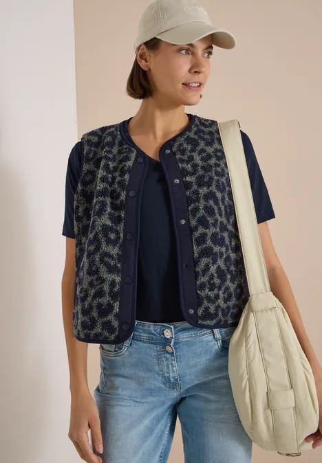 Frau mit weißem Cap, dunkelblauer Bluse, gemusterter Weste, Jeans und cremefarbener Stepp-Tasche.
