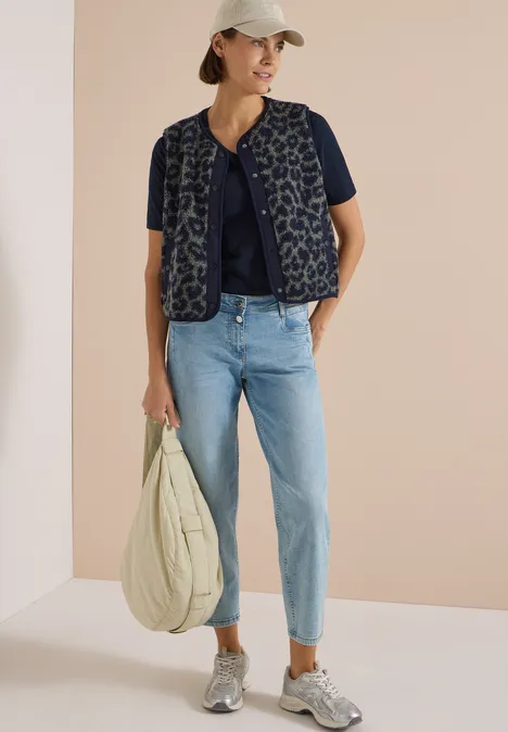 Frau in weißer Kappe trägt gestepste Weste über dunkelblauem Shirt, hellblaue Jeans, graue Sneaker und große Tasche.