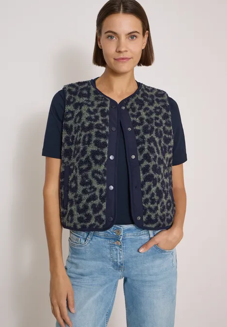 Frau in blauem T-Shirt mit grüner, leo-gemusterter Weste und hellen Jeans vor weißem Hintergrund.