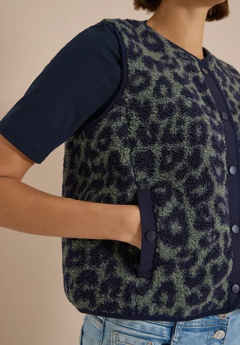 Eine Person trägt eine dunkelgrüne, leopardgemusterte Weste mit Knöpfen über einem marineblauen T-Shirt und Jeans.