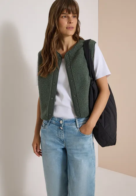 Frau in weißem T-Shirt, Jeans, grüner Teddy-Weste mit Kapuze und schwarzer Schultertasche.