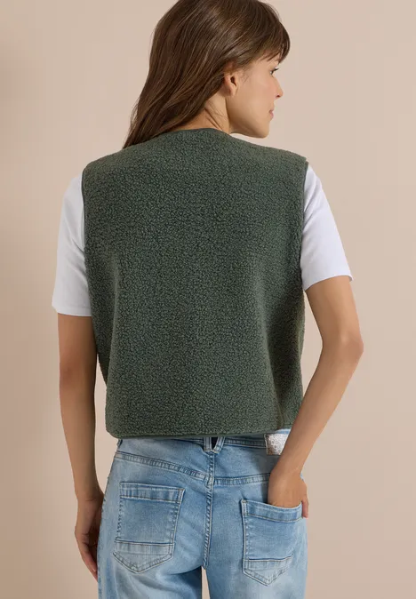 Eine Frau in graugrüner Fleece-Weste, weißem T-Shirt und Jeans, von hinten.