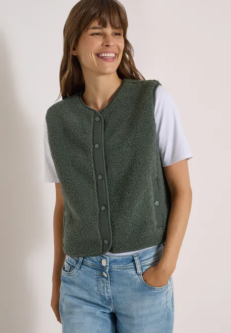 Eine Frau in grüner, geknöpfter Fleece-Weste, weißem T-Shirt und Jeans lächelt mit einer Hand in der Hosentasche.