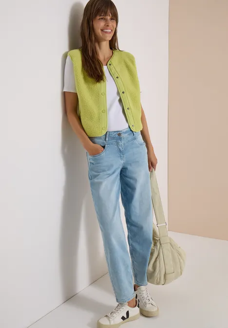 Junge Frau lehnt lässig an der Wand, trägt Jeans, weißes T-Shirt und grüne Weste, dazu schlichte Sneaker und Tasche.