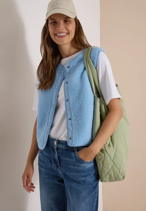 Lächelnde Frau mit weißem T-Shirt, hellblauer Strickweste, Jeans, grüner Tasche und beigem Cap.