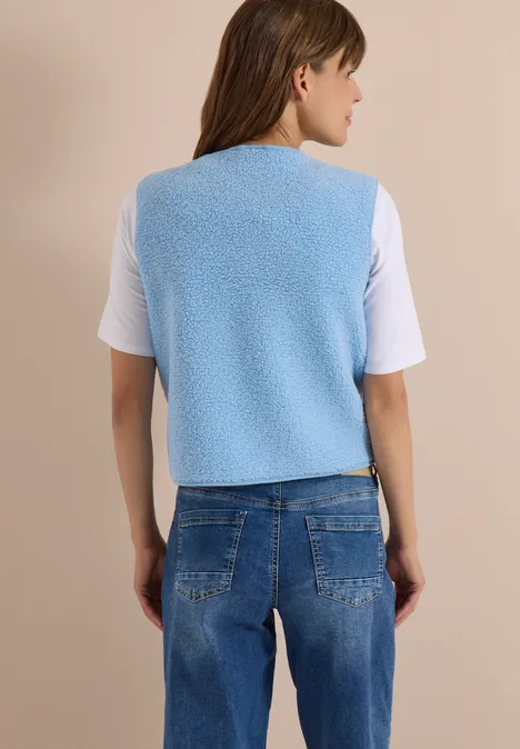 Frau von hinten in hellblauer Teddy-Weste, weißem T-Shirt und blauen Jeans vor beigem Hintergrund.