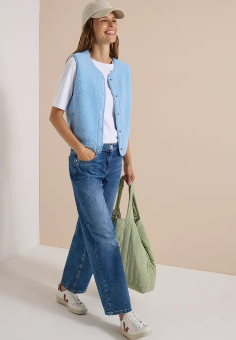 Frau in weißem T-Shirt, hellblauer Weste, Jeans und Cap trägt grüne Tasche und läuft lächelnd.