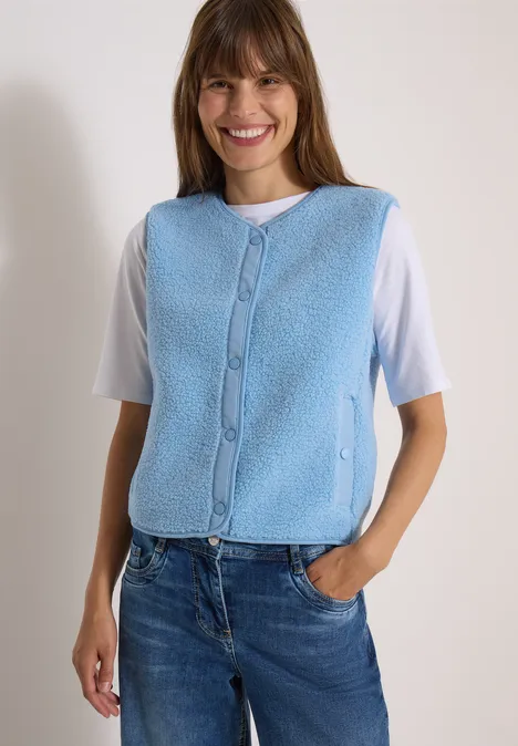 Frau trägt hellblaue Fleece-Weste über weißem Shirt und Jeans, lächelt vor neutralem Hintergrund.