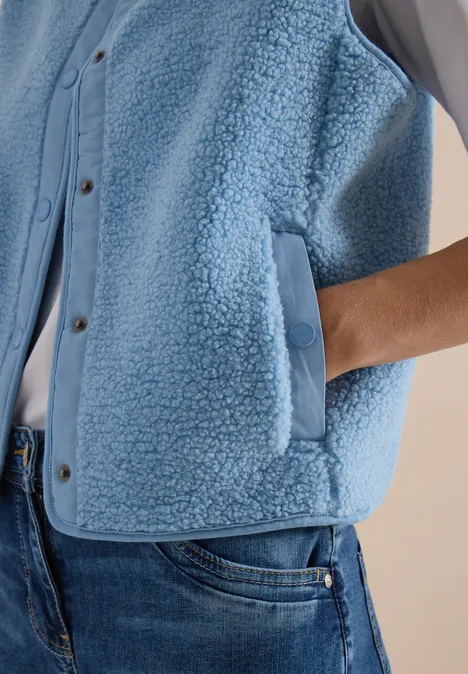 Flauschige hellblaue Weste mit Taschen, eine Hand ist in der rechten Tasche.