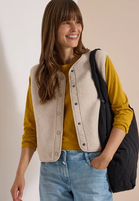 Teddy-Weste mit Softshelldetails teddy vanilla white Frau mit gelbem Pullover, beiger Teddy-Weste, Jeans und schwarzer Umhängetasche lächelt.