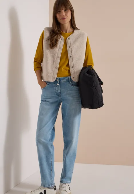 Teddy-Weste mit Softshelldetails teddy vanilla white Frau in senfgelbem Pullover, beigem Gilet und blauen Jeans, trägt eine dunkle Tasche vor hellem Hintergrund.
