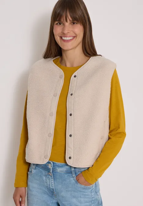 Teddy-Weste mit Softshelldetails teddy vanilla white Lächelnde Frau trägt gelbes Shirt und beige Weste, lässig in Jeans, Hände in Tasche.