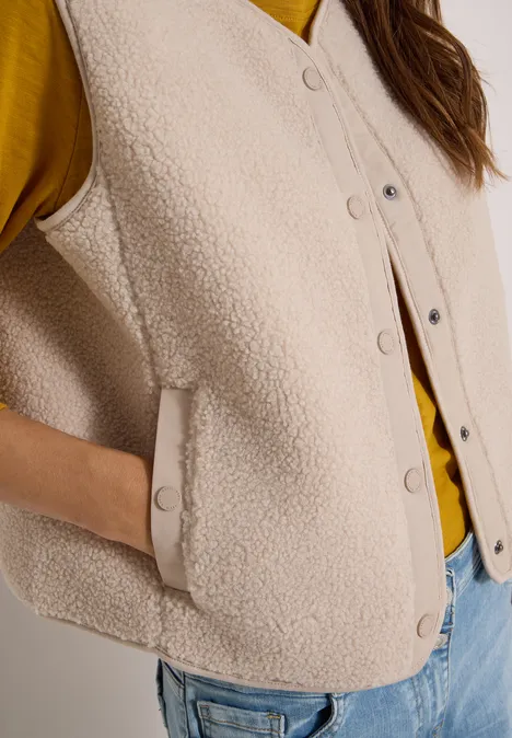 Teddy-Weste mit Softshelldetails teddy vanilla white Eine Nahaufnahme einer beigen Teddy-Fleece-Weste mit Druckknöpfen und Eingrifftasche über einem gelben Pullover.