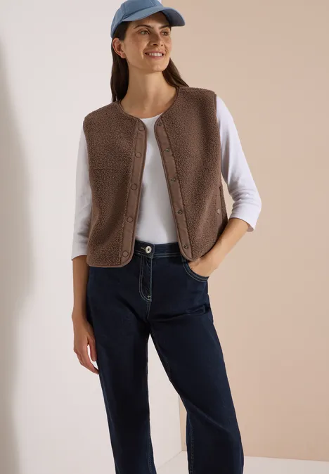 Eine junge Frau trägt eine braune Fleece-Weste, weißes Langarmshirt, blaue Jeans und eine blaue Baseballkappe.