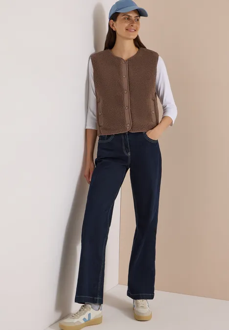 Eine Frau lehnt an der Wand in brauner Weste, weißem Shirt, dunklen Jeans, Cap und Sneakers.