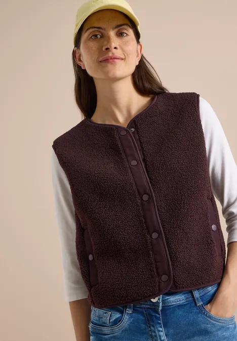 Frau mit dunkelbrauner Fleeceweste, weißem Langarmshirt, Jeans und beiger Mütze.