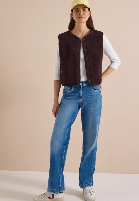 Frau mit gelber Mütze, braunem ärmellosem Weste, weißem 3/4-Shirt, blauen Jeans und weißen Sneakern vor neutralem Hintergrund.