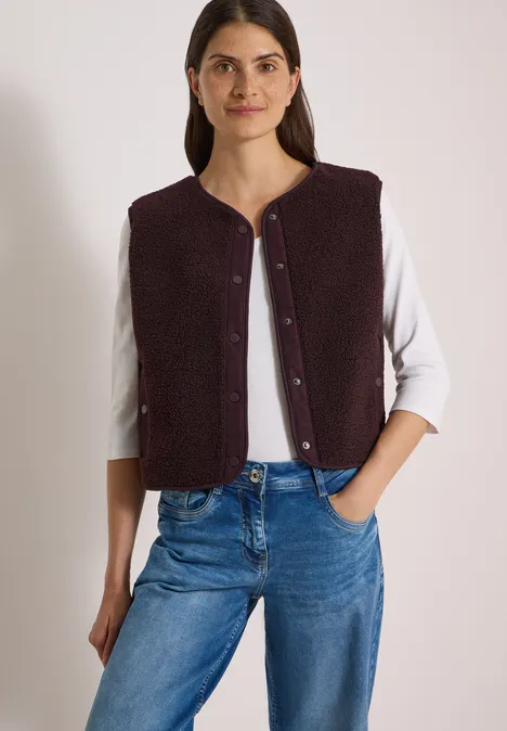 Frau in weißem Langarmshirt, violetter Weste und Jeans, Hände in den Taschen.