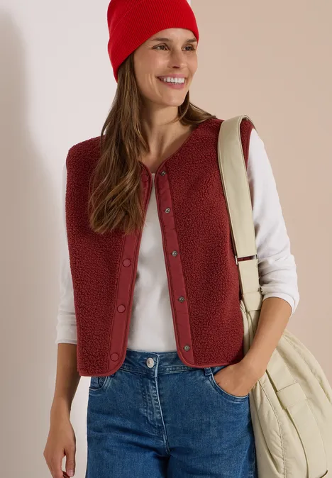 Teddy-Weste mit Softshelldetails russet red Eine junge Frau trägt rote Weste und Mütze, ein weißes Shirt, Jeans und eine Cremetasche, sie lächelt.