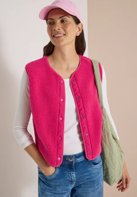 Frau in pinker Weste, weißem Shirt, Jeans und Kappe mit grünem Shopper, lächelnd seitlich blickend.